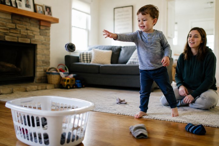 A 3-year-old tossing a rolled sock toward a laundry basket while a parent watches