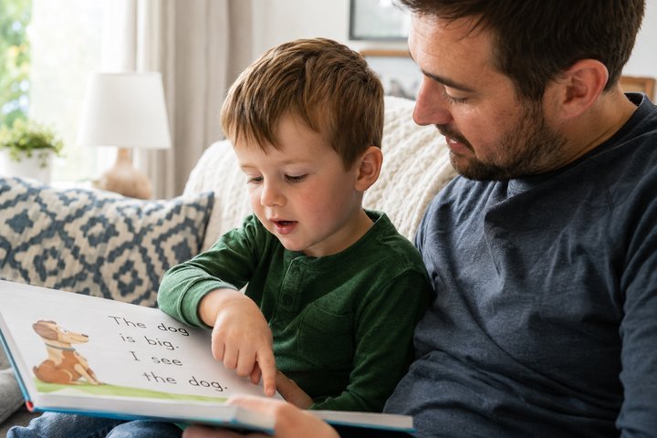 A young child pointing at and sounding out a word in an early reader while a parent sits beside them